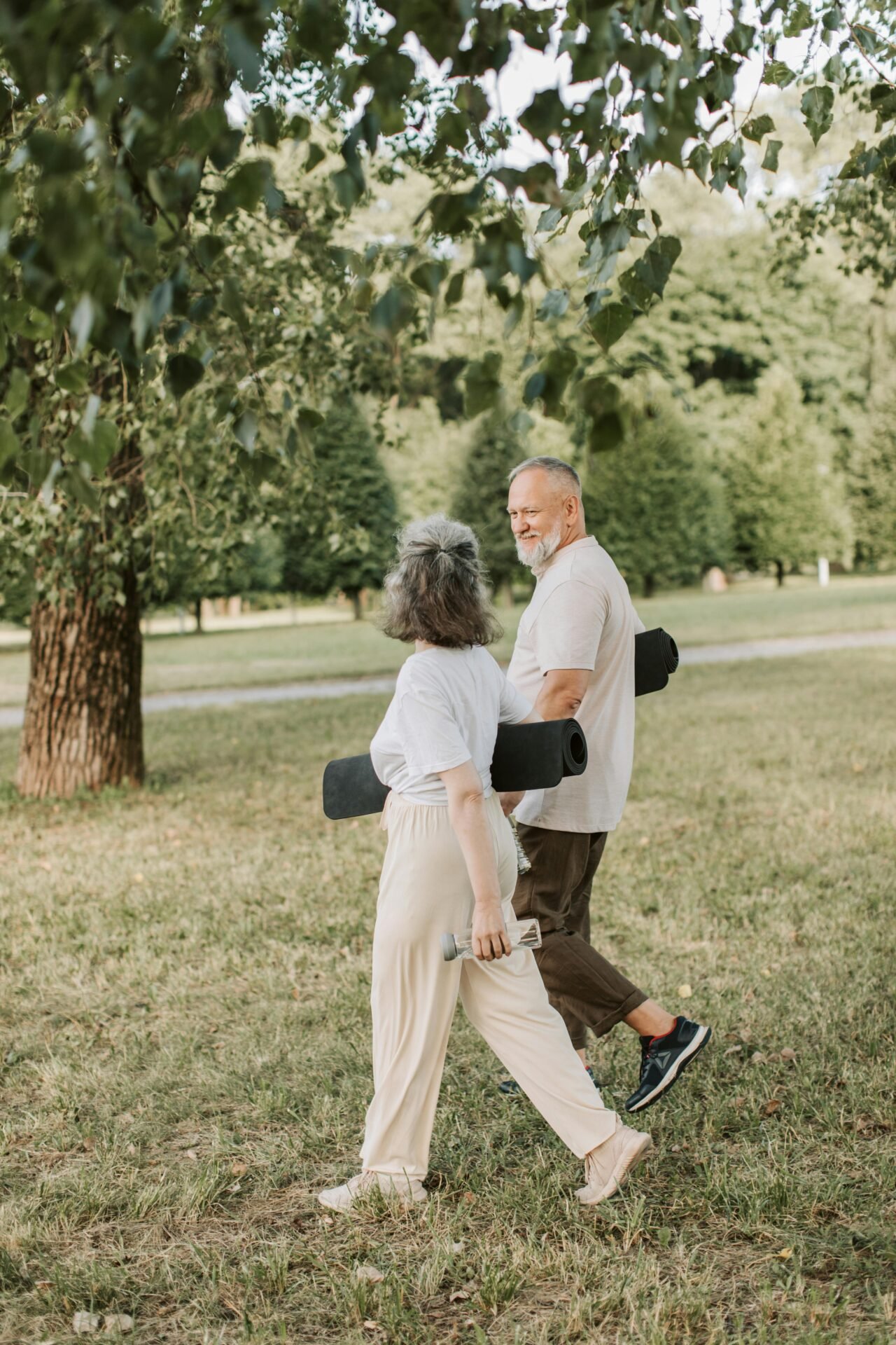 Elderly couple carrying yoga mats and water bottles, walking in a sunny park for wellness.